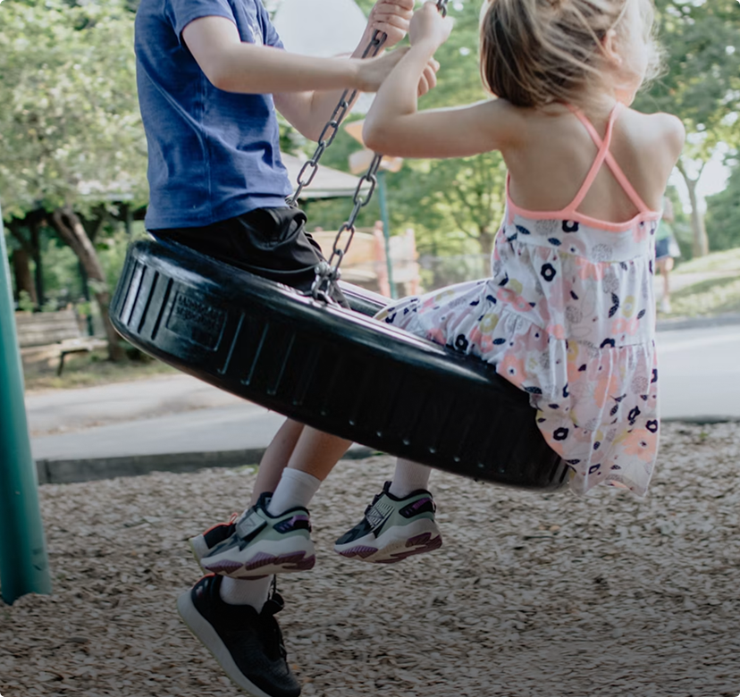 Two children on a tire swing at a playground, one in a blue shirt and one in a floral dress.
