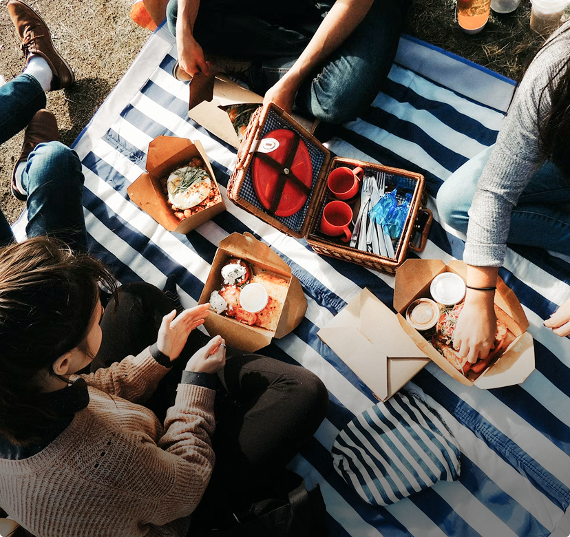 People sitting on a striped blanket sharing food from takeout boxes outdoors.