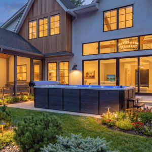 Modern house patio at dusk with lit windows, outdoor seating, and a large hot tub in the yard