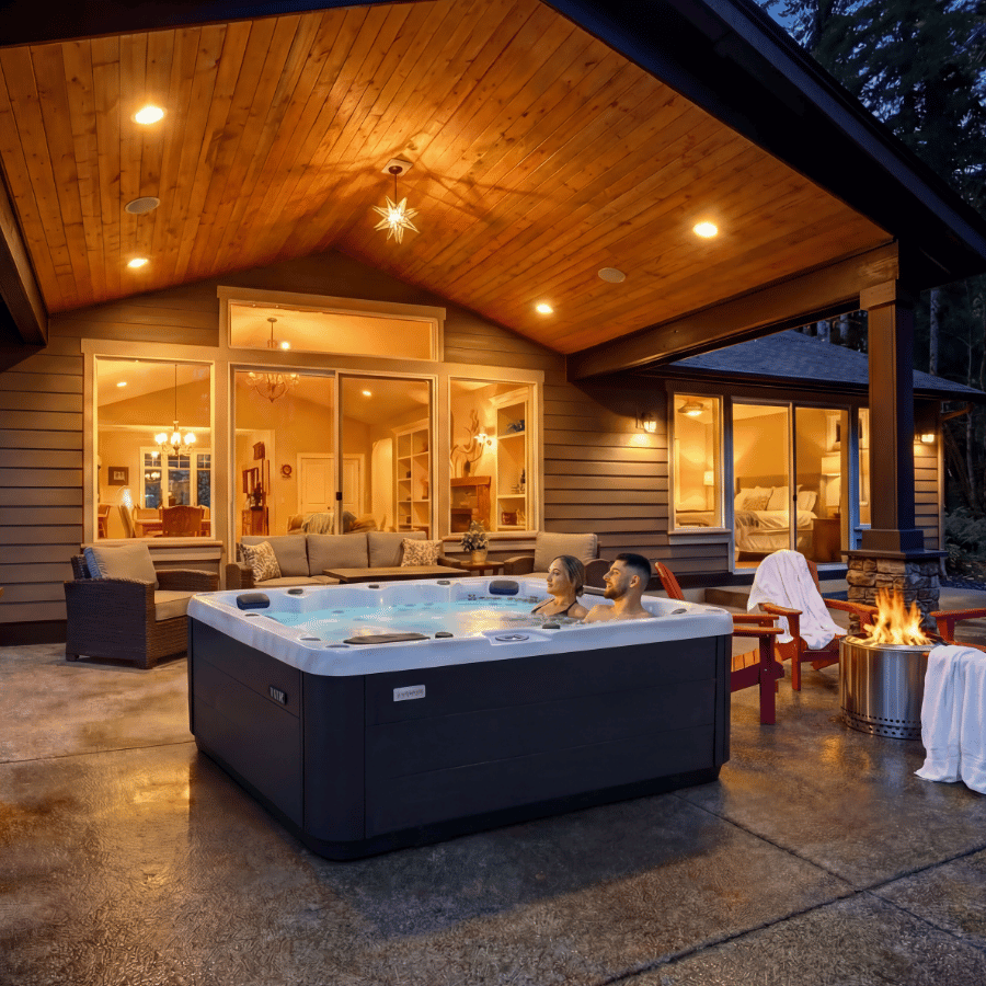 Warm-lit patio with a hot tub, lounge seating, and a fire pit outside a modern cabin at dusk