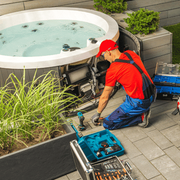 Technician in red uniform repairing hot tub plumbing beside a patio spa with tools nearby