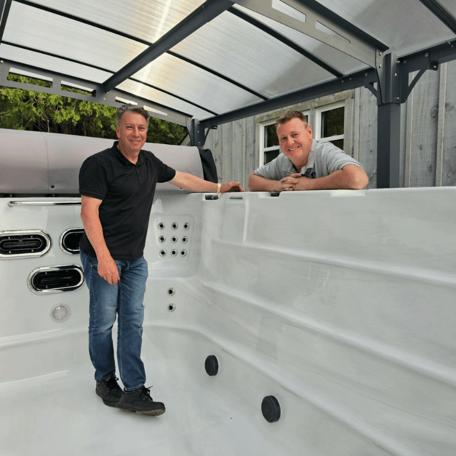 Two men standing in a white hot tub shell under a covered outdoor patio, smiling at the camera.