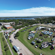 Aerial view of a grassy fairground with tents, parked cars, and a lake in the background.