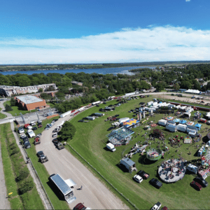 Aerial view of a grassy fairground with tents, parked cars, and a lake in the background.