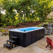 Black hot tub on a stone patio beside a waterfall and trees, with red chairs nearby