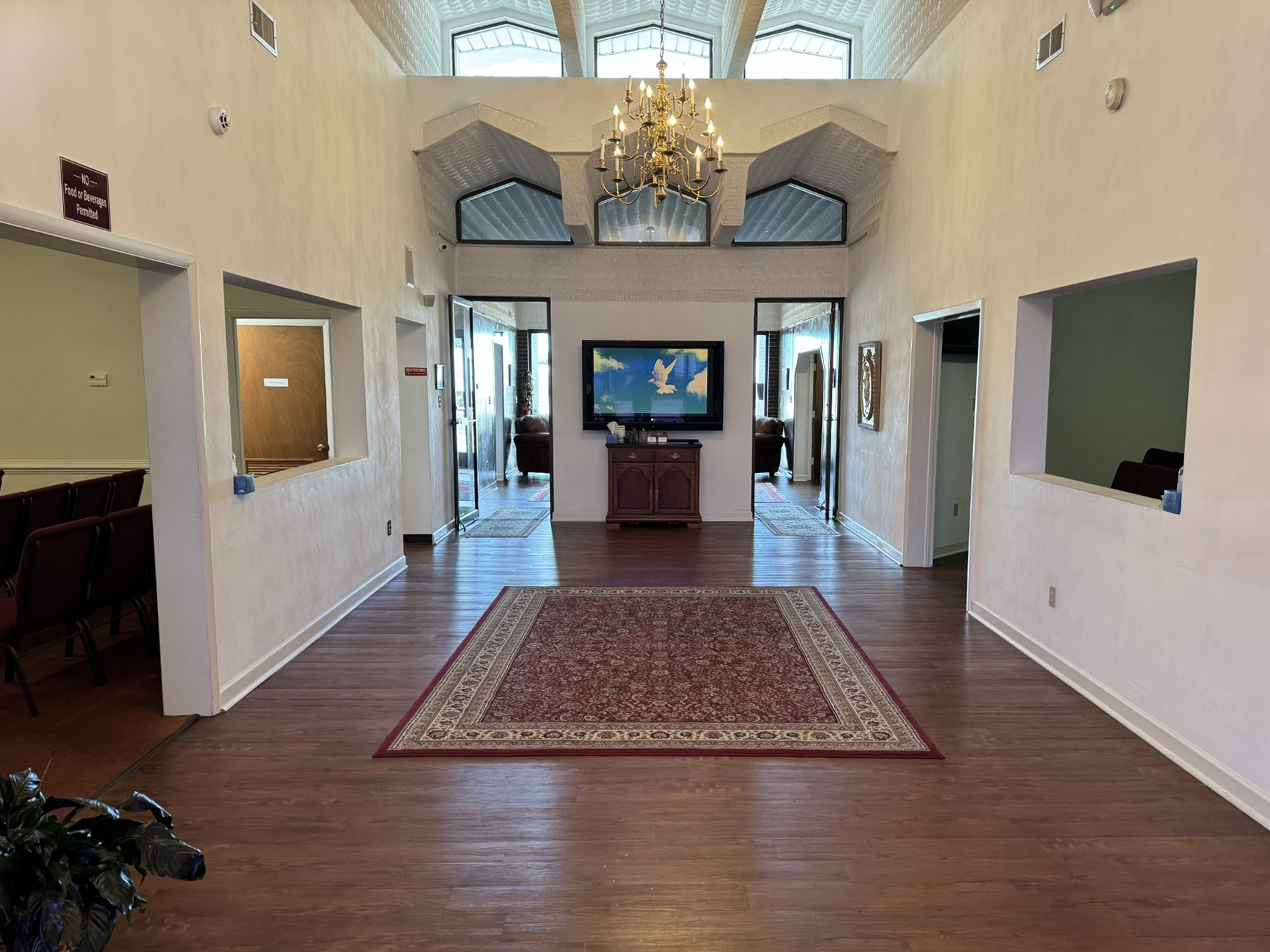 Spacious room with a rug, TV, and arched windows. Doors on each side, a chandelier, and wooden floors.