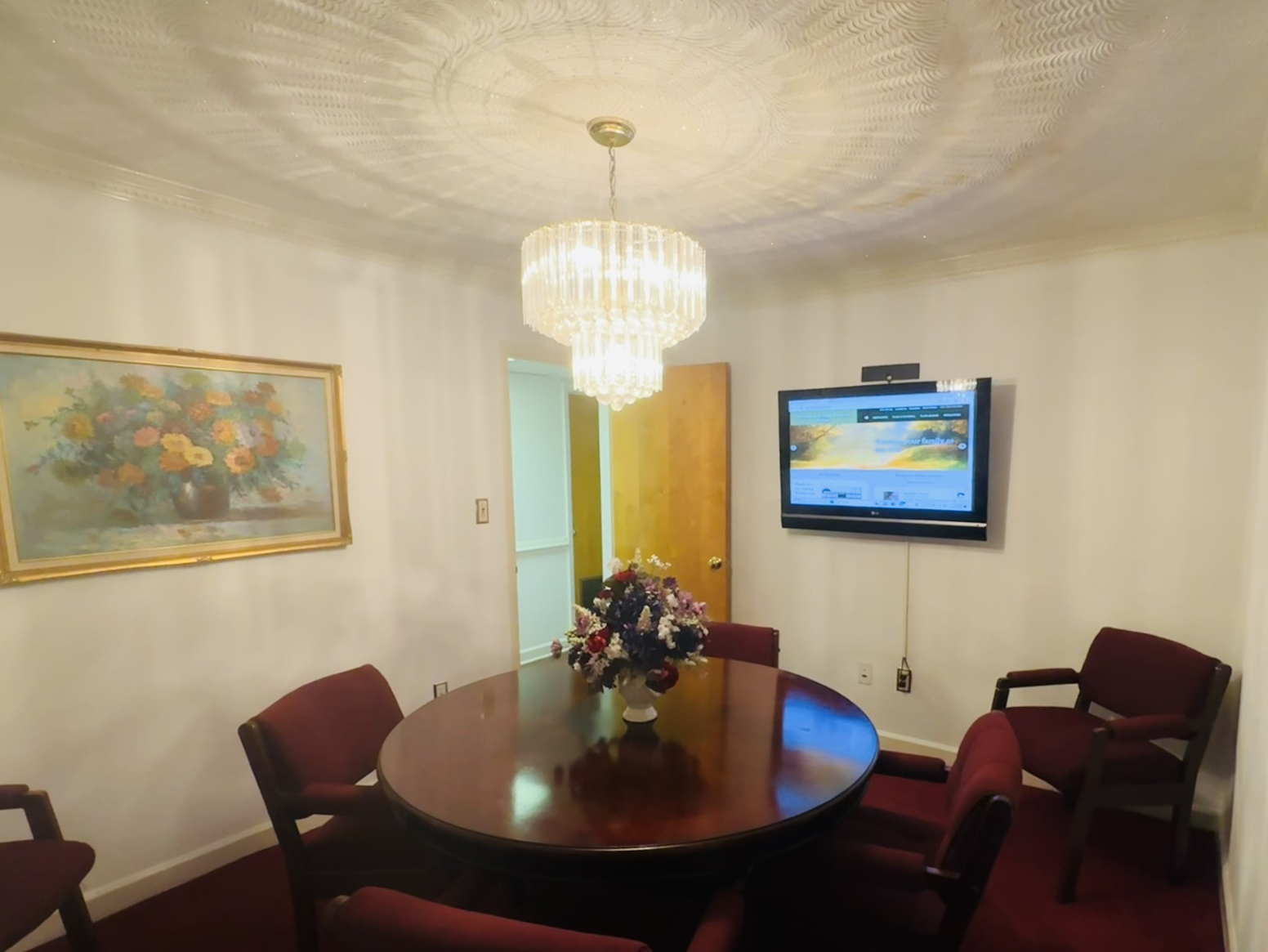 Small room with a dark wood table, chairs, chandelier, TV, and floral arrangement; a painting hangs on the wall.