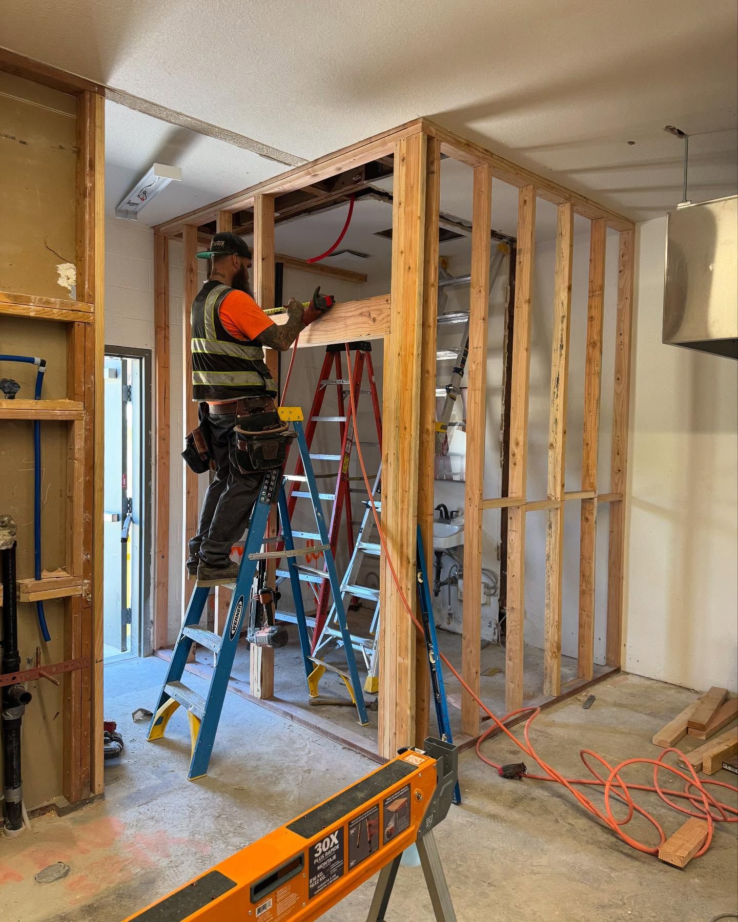 Construction worker framing interior wall, wearing safety vest, standing on ladder. New construction in process.