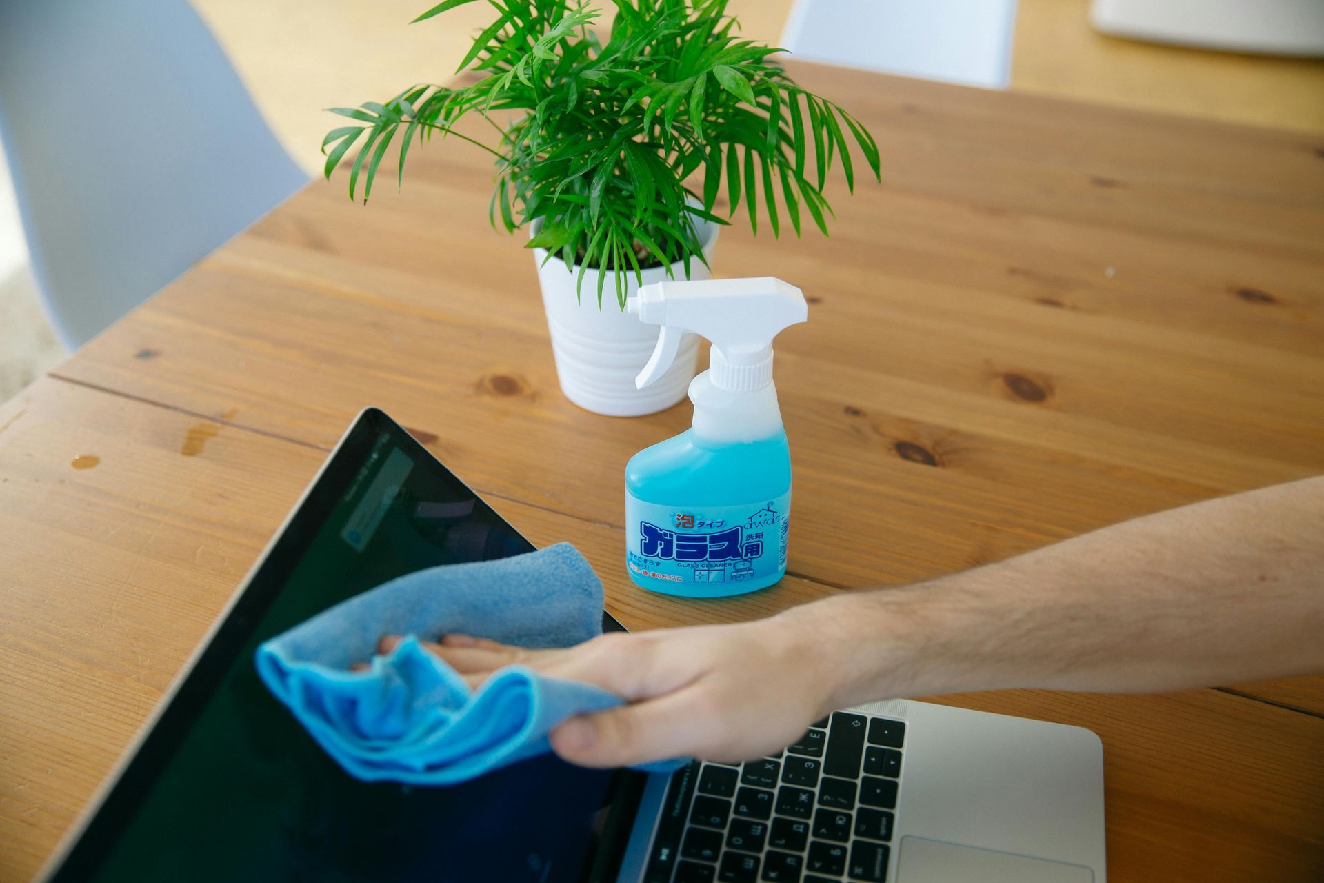 Hand cleaning a laptop with blue cloth and spray bottle on a wooden table near a potted plant