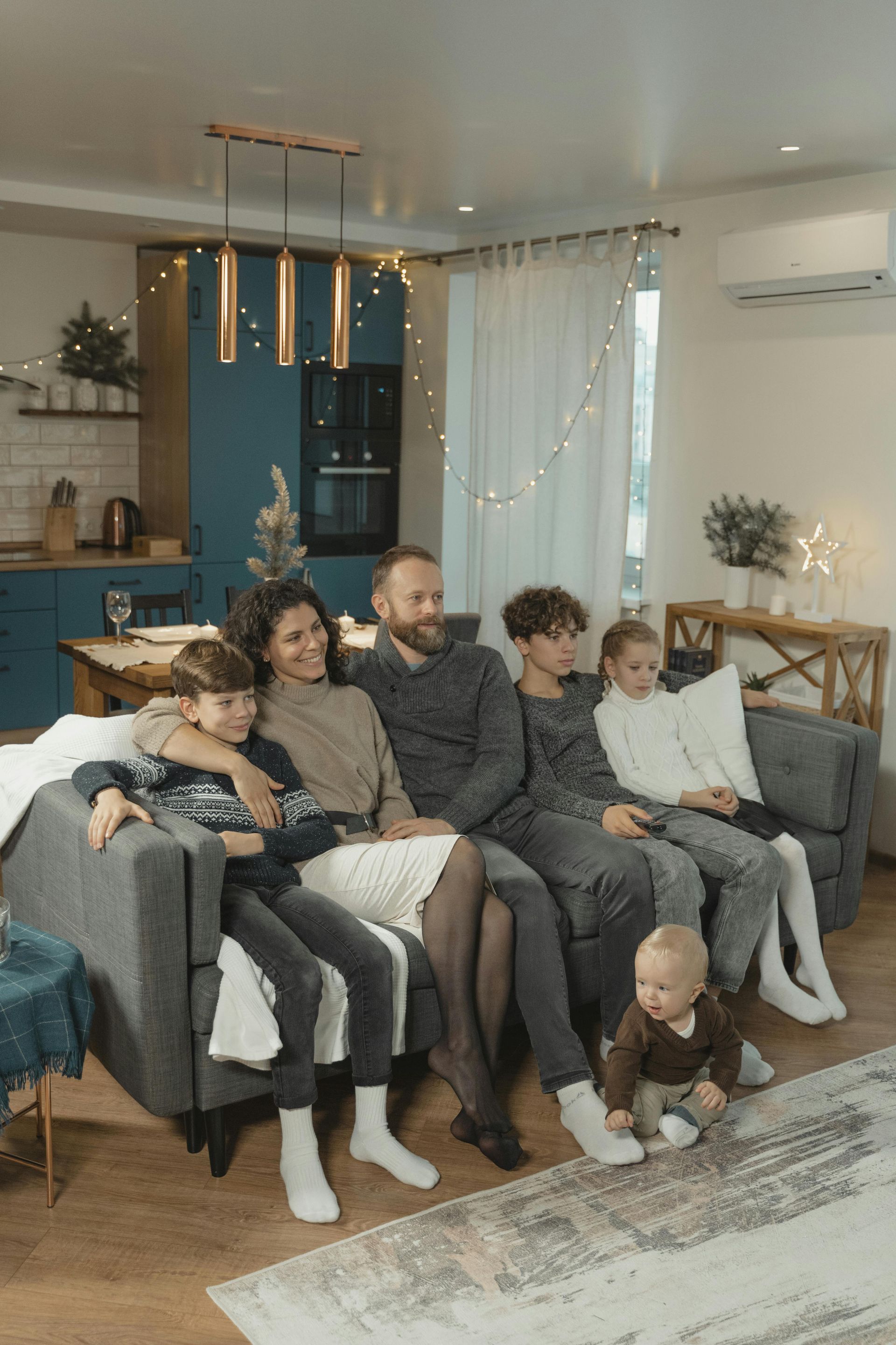Family sitting together on a gray couch in a cozy living room, smiling and relaxed, with a baby on the floor.