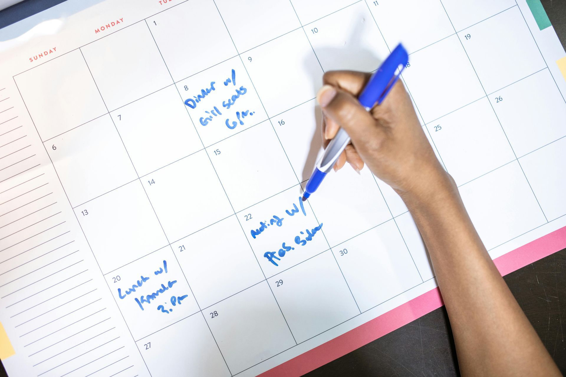 A hand uses a blue pen to write on a paper monthly calendar, which features handwritten notes in several squares.