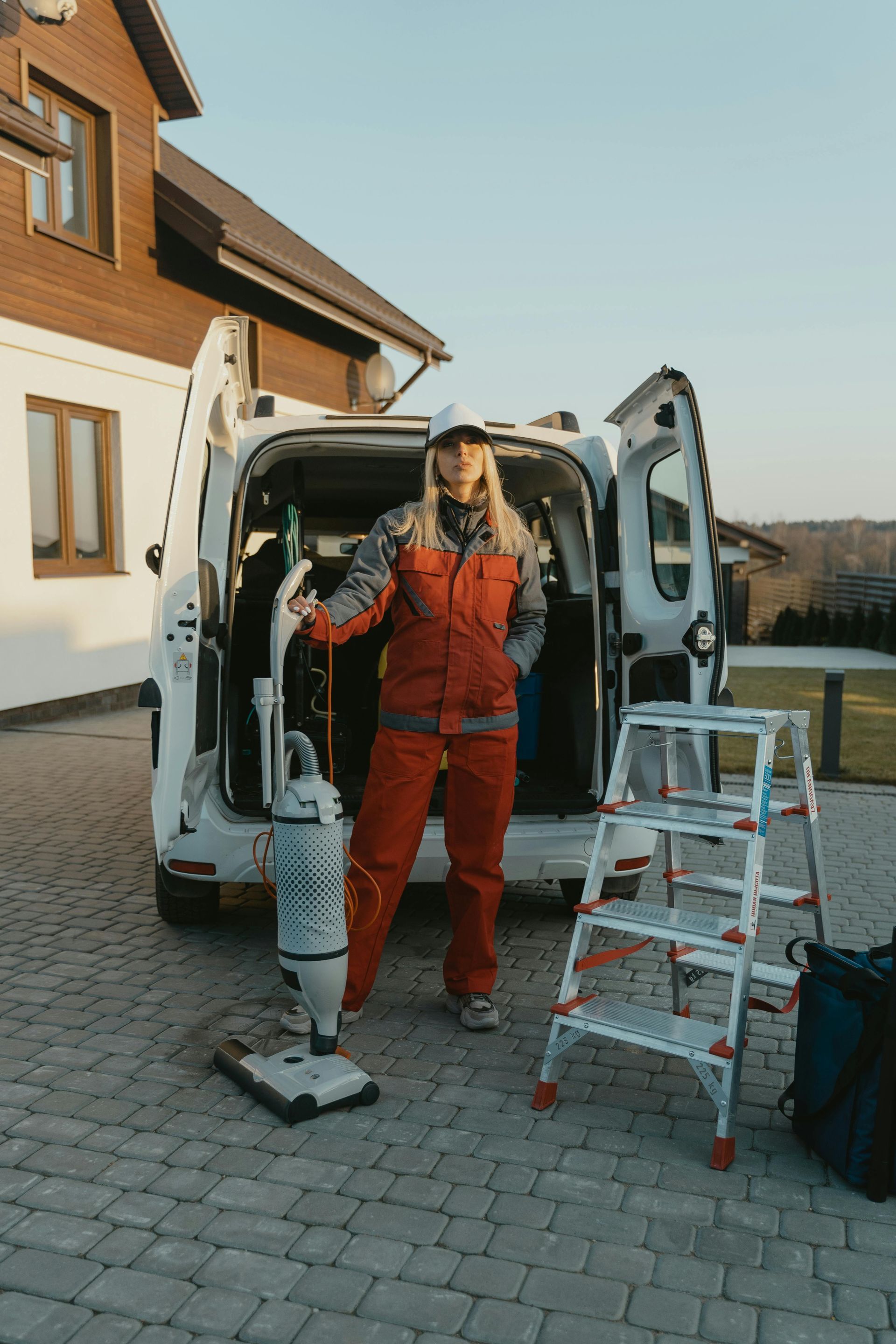 A person in orange work clothes stands before a van with an open door, holding a vacuum cleaner next to a step ladder.