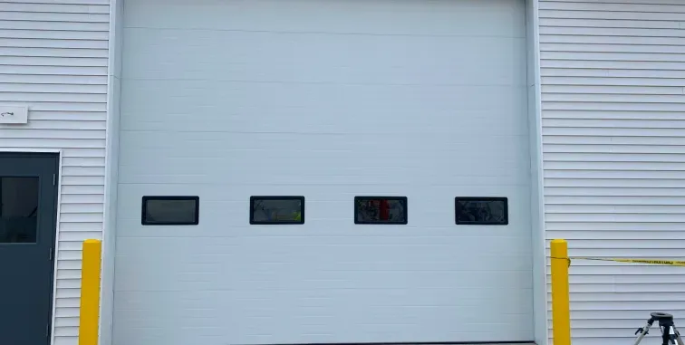 Industrial building facade with a large white roll-up garage door and yellow safety bollards.