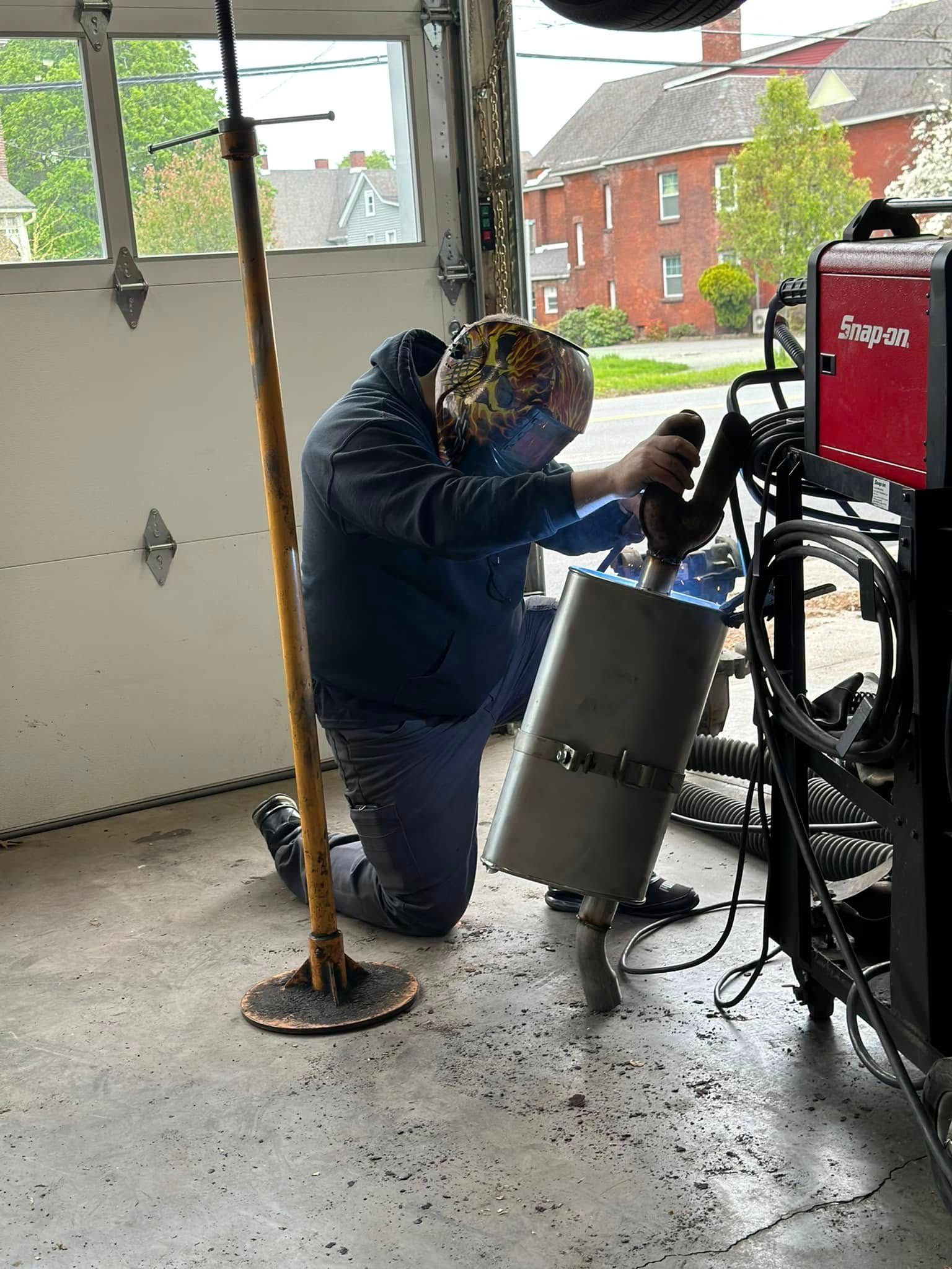Person welding a metal cylinder in a garage beside a red welding machine. | 1st Class Alignment & Repair