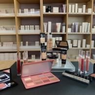 Makeup display on black table with pink palette, lip gloss, and shelves of beauty products in background