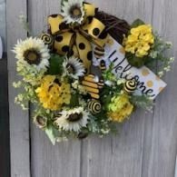 Yellow and white flower wreath with black bee accents and a “Welcome” sign on a gray wooden door