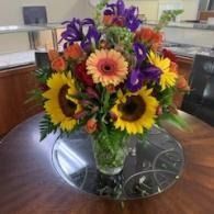 Colorful bouquet of sunflowers, daisies, and purple flowers in a vase on a glass-topped table