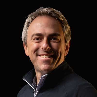 A man in a black jacket is smiling in front of a black background.