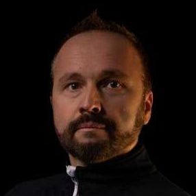 A man with a beard is wearing a black jacket and looking at the camera.