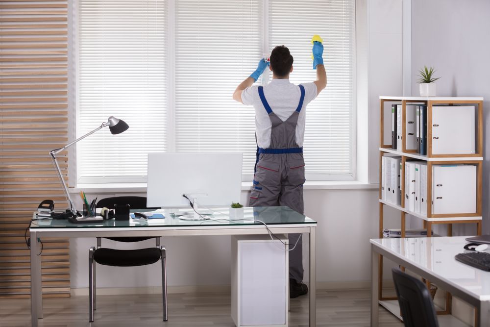 Person cleaning window blinds in a well-lit office, wearing blue gloves and overalls.