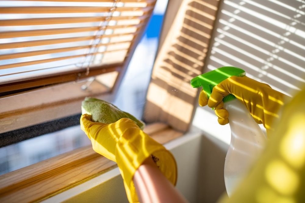 Hands in yellow gloves cleaning blinds with a spray bottle and sponge near a window.