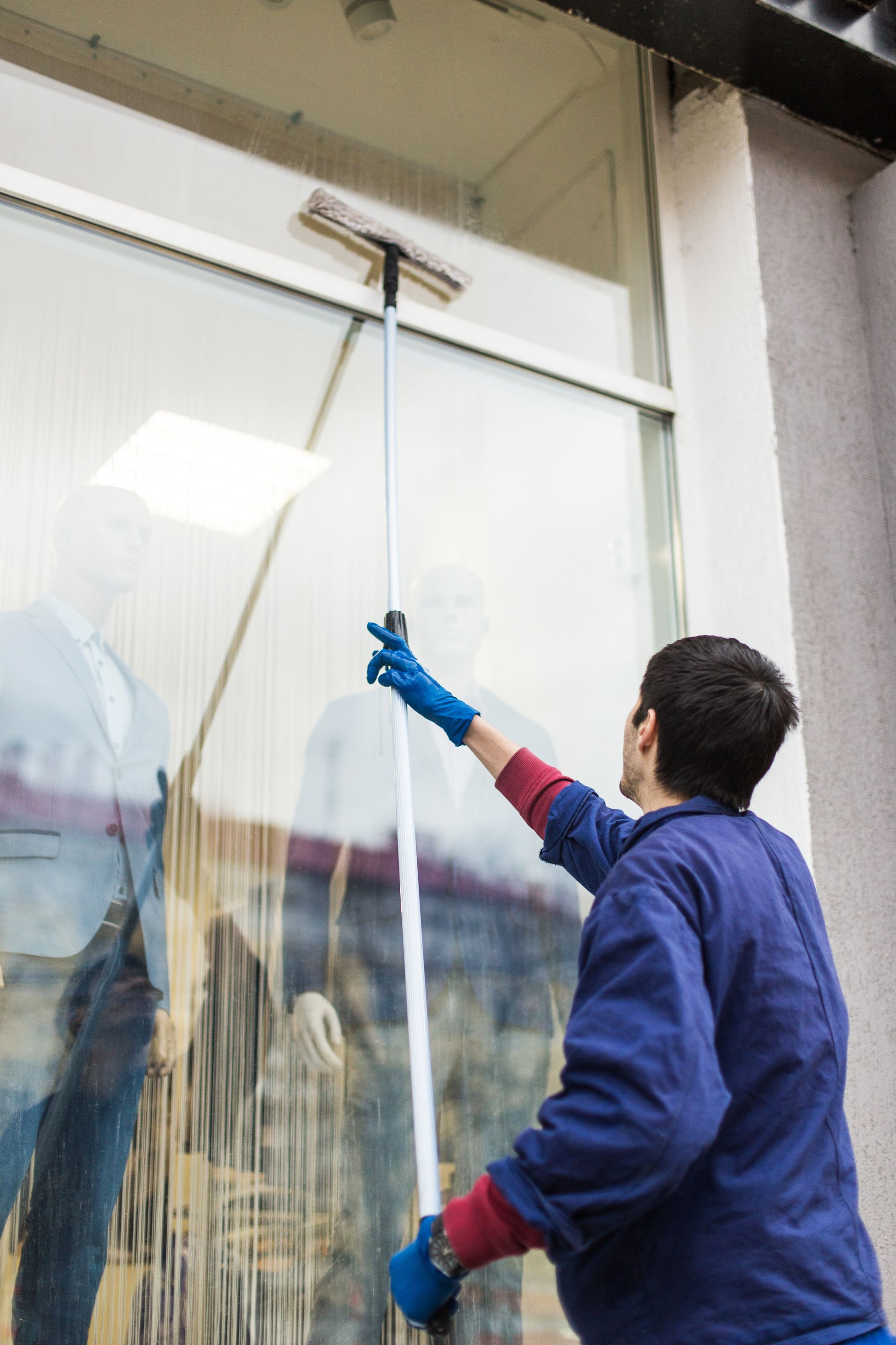 Person in blue overalls cleaning a window with a squeegee on a pole outdoors.