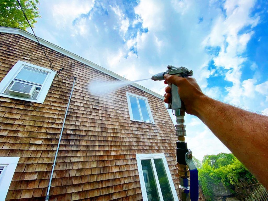 Person spraying a brown shingled house with a water gun under a blue sky.