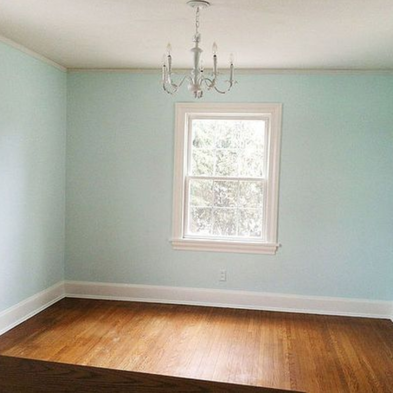 Empty room with light blue walls, hardwood floor, white trim, window, and chandelier.