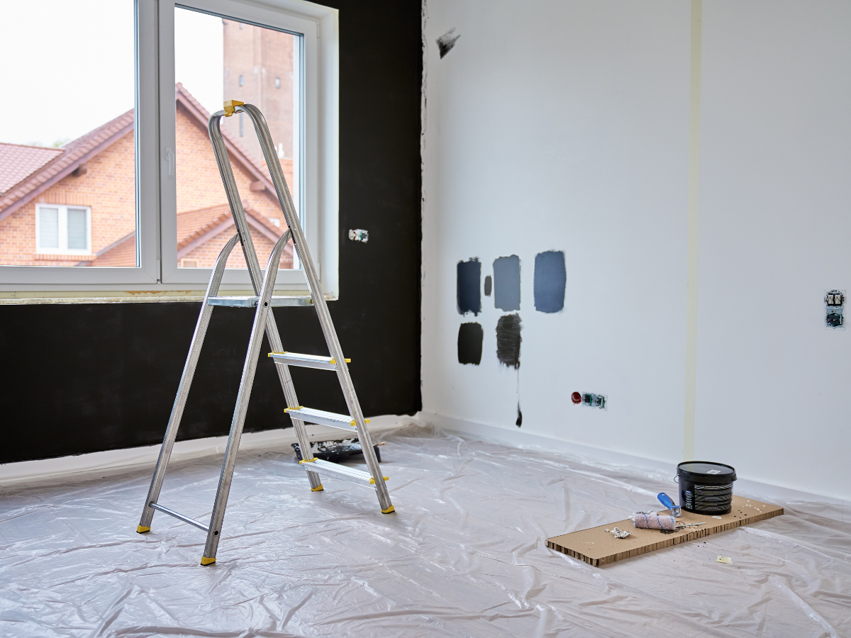 Empty room under renovation with a step ladder, paint swatches on the wall, and supplies on the floor