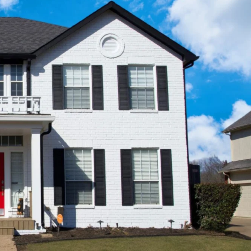 White two-story house with black shutters, a red front door, and a driveway under a blue sky