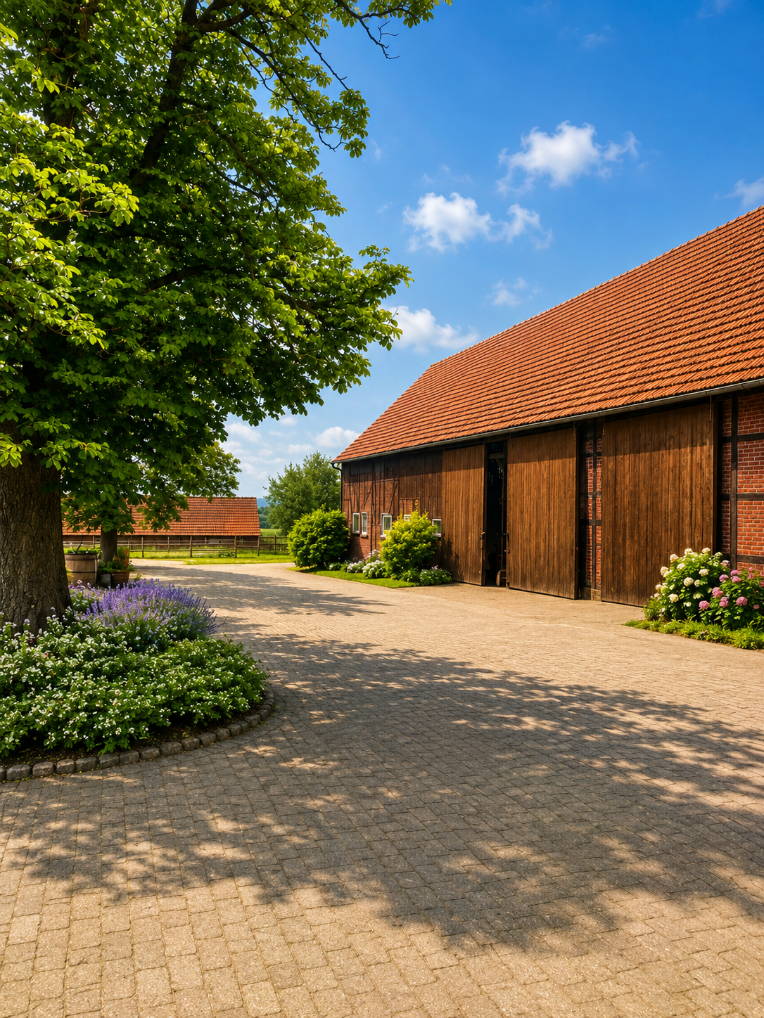 Kieshof neben einer Scheune mit roten Ziegeln, einem belaubten Baum, Blumenbeeten und blauem Himmel.