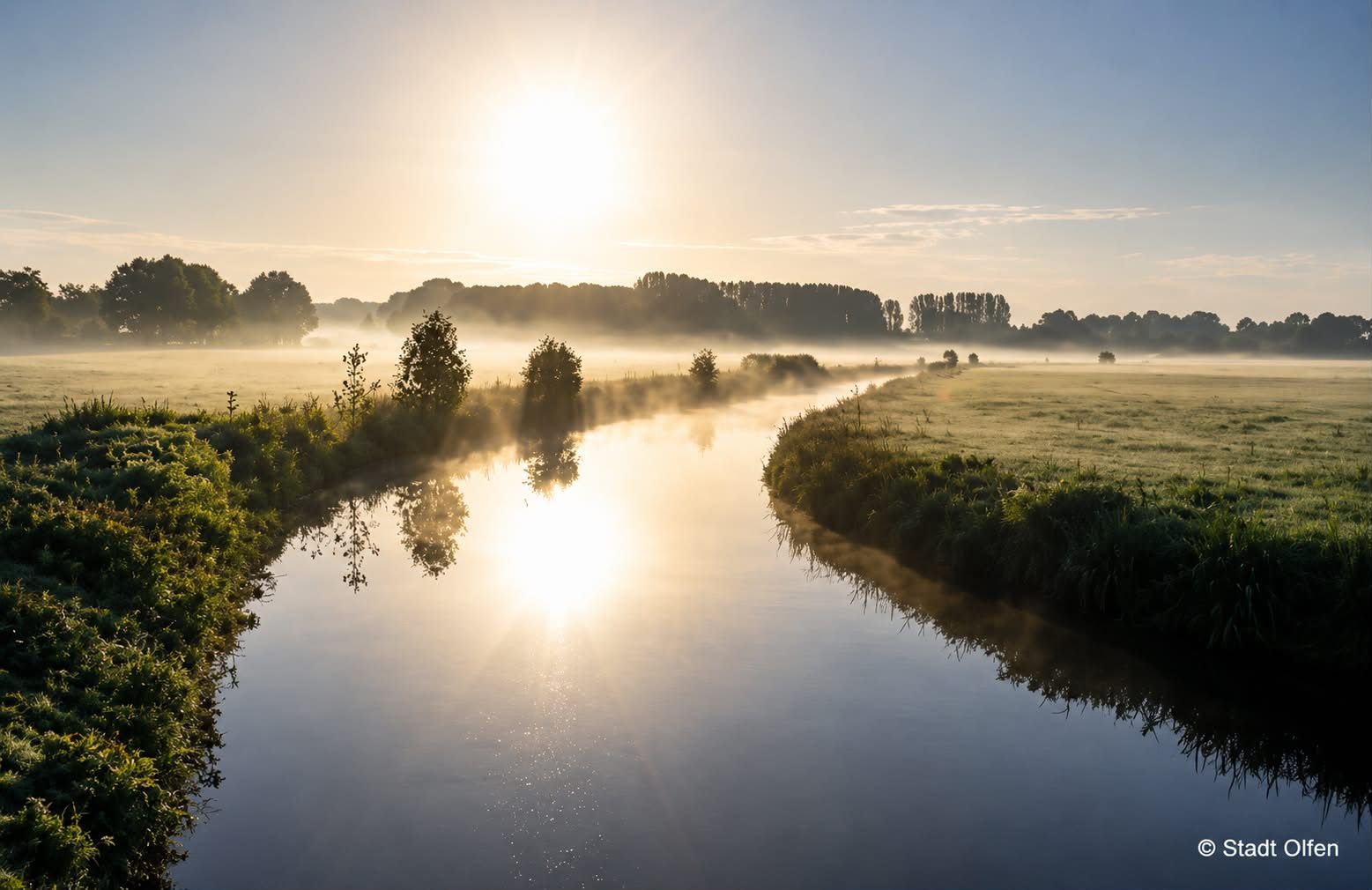 Sonnenaufgang über einem nebligen Fluss, der sich durch Felder schlängelt mit Bäumen