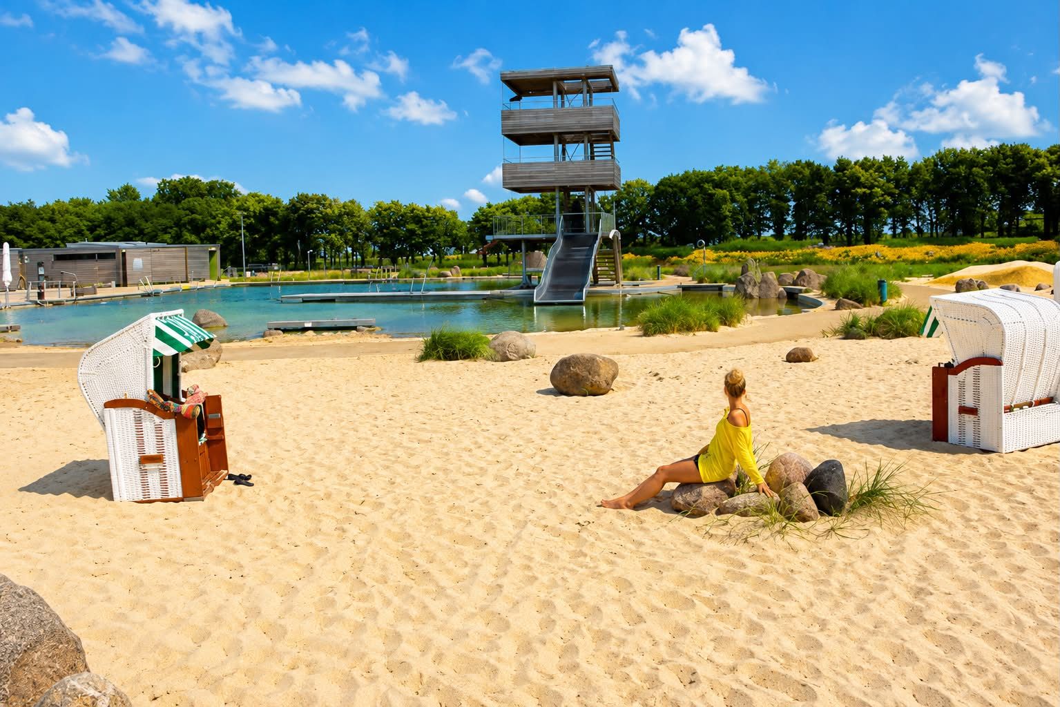 Sonniger Strand mit Sand, verstreuten Felsen, zwei weißen Stühlen und einem hohen Sprungturm am Wasser