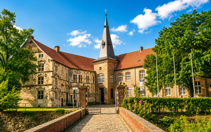 Historischer Innenhof mit Gebäuden mit roten Ziegeldächern und einem hohen Kirchturm unter blauem Himmel