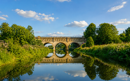 Steinbrücke mit drei Bögen über einem ruhigen Fluss, in dem sich grüne Bäume und ein blauer Himmel mit Wolken spiegeln.