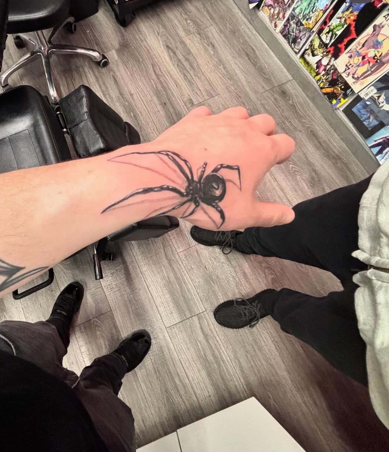 Hand with large black spider tattoo hovering over a gray store floor, surrounded by people’s shoes.