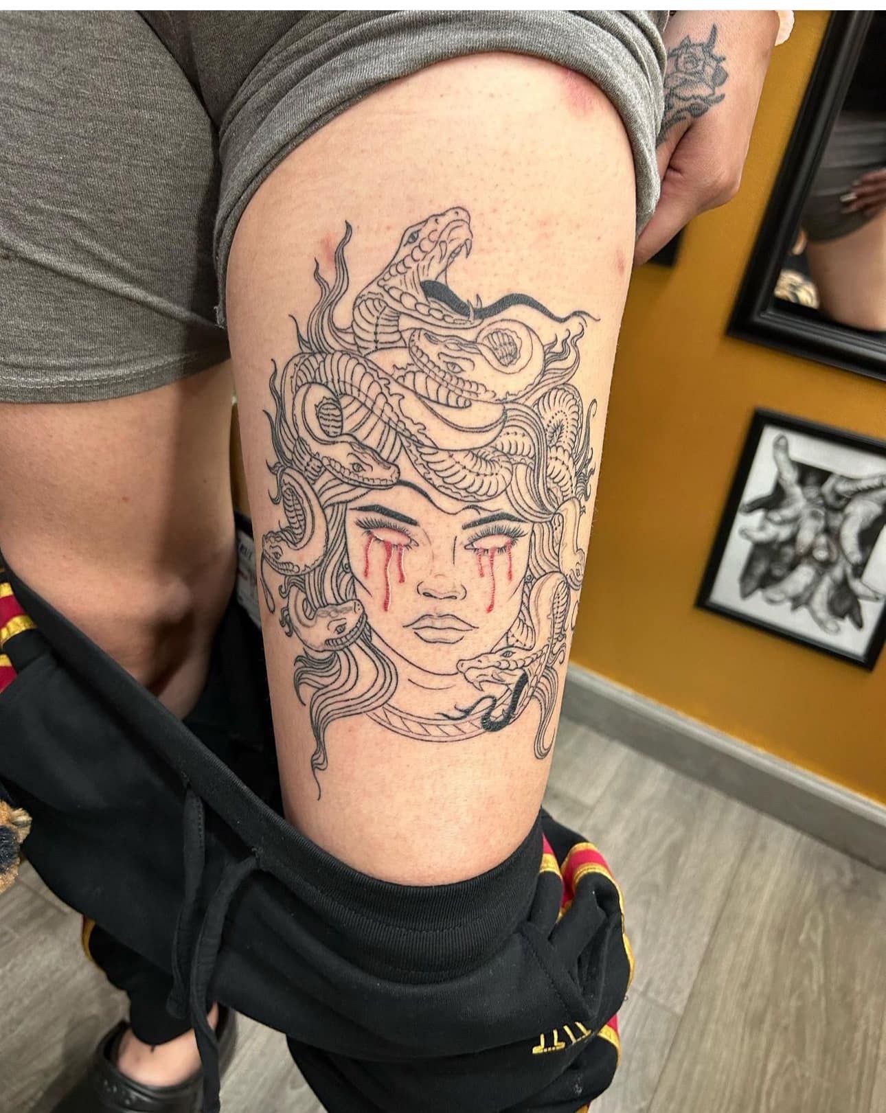 Close-up of a black-and-red tattoo on a thigh, showing a detailed mythical face design.