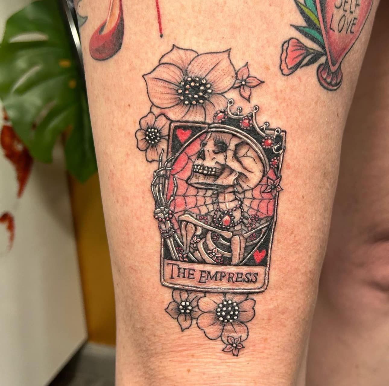 Tarot card “The Empress” tattoo in black, red, and pink on a person’s leg, with floral accents
