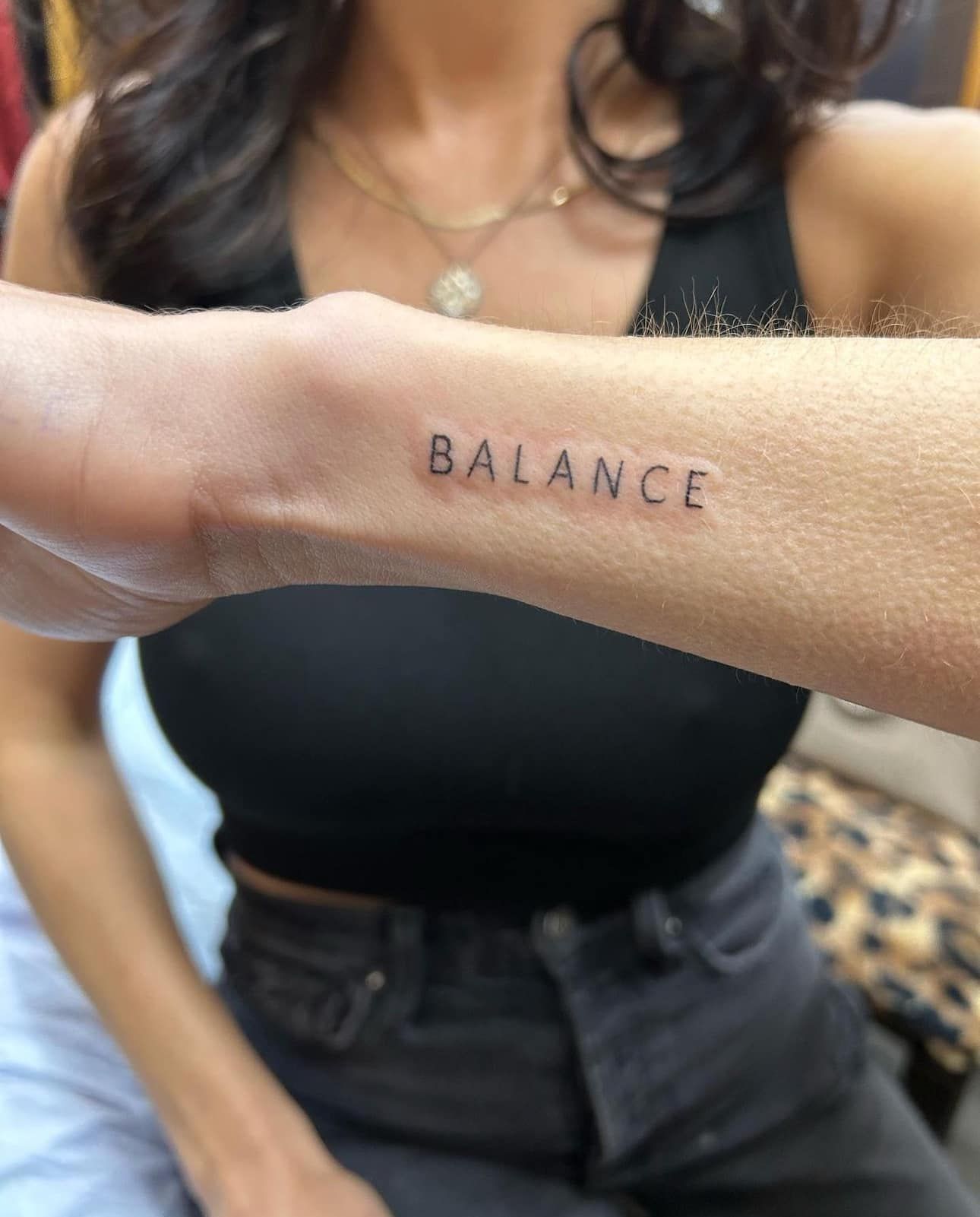 Forearm with small “BALANCE” tattoo, held in front of a person wearing a black top and jeans.