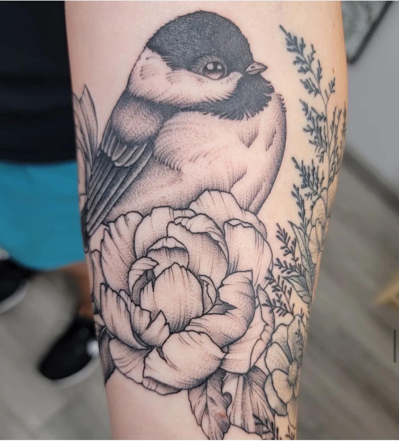 Black-and-gray tattoo of a bird perched on a large flower with leafy stems on skin