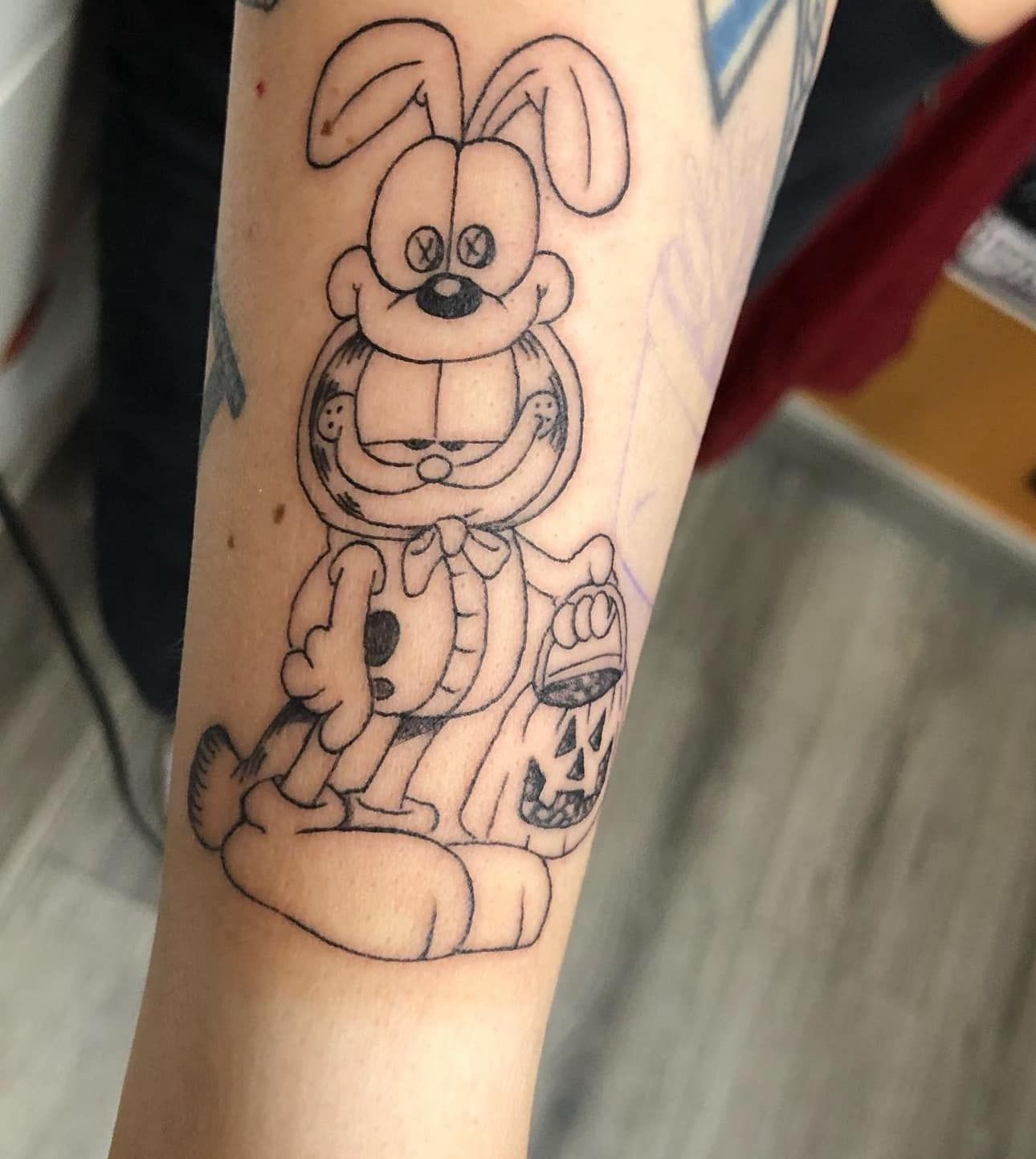 Black-and-white cartoon rabbit tattoo on a person’s forearm, holding a bottle and standing beside a small figure