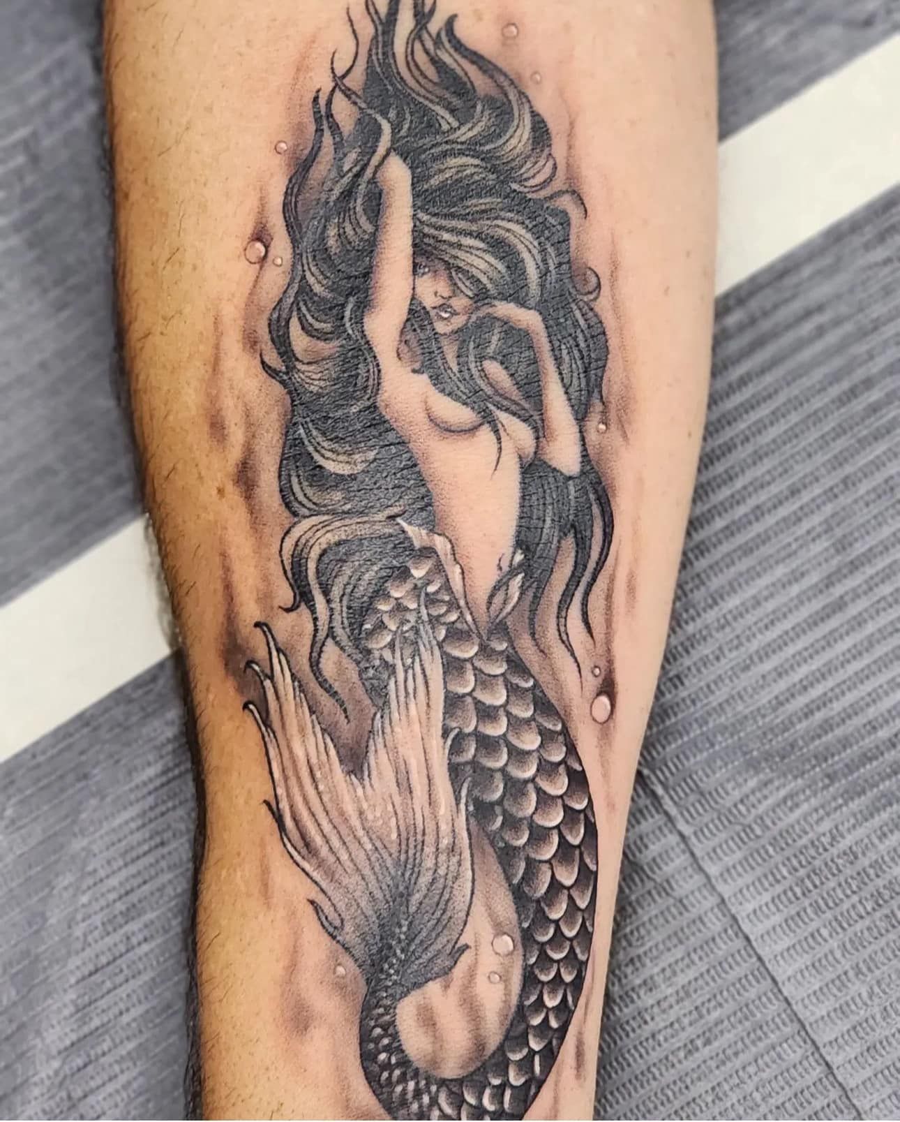 Black-and-gray mermaid tattoo on a forearm, with flowing hair and detailed scales.