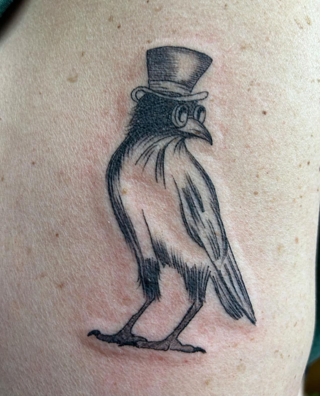 Small black-and-gray tattoo of a bird wearing a top hat and goggles, standing in profile.