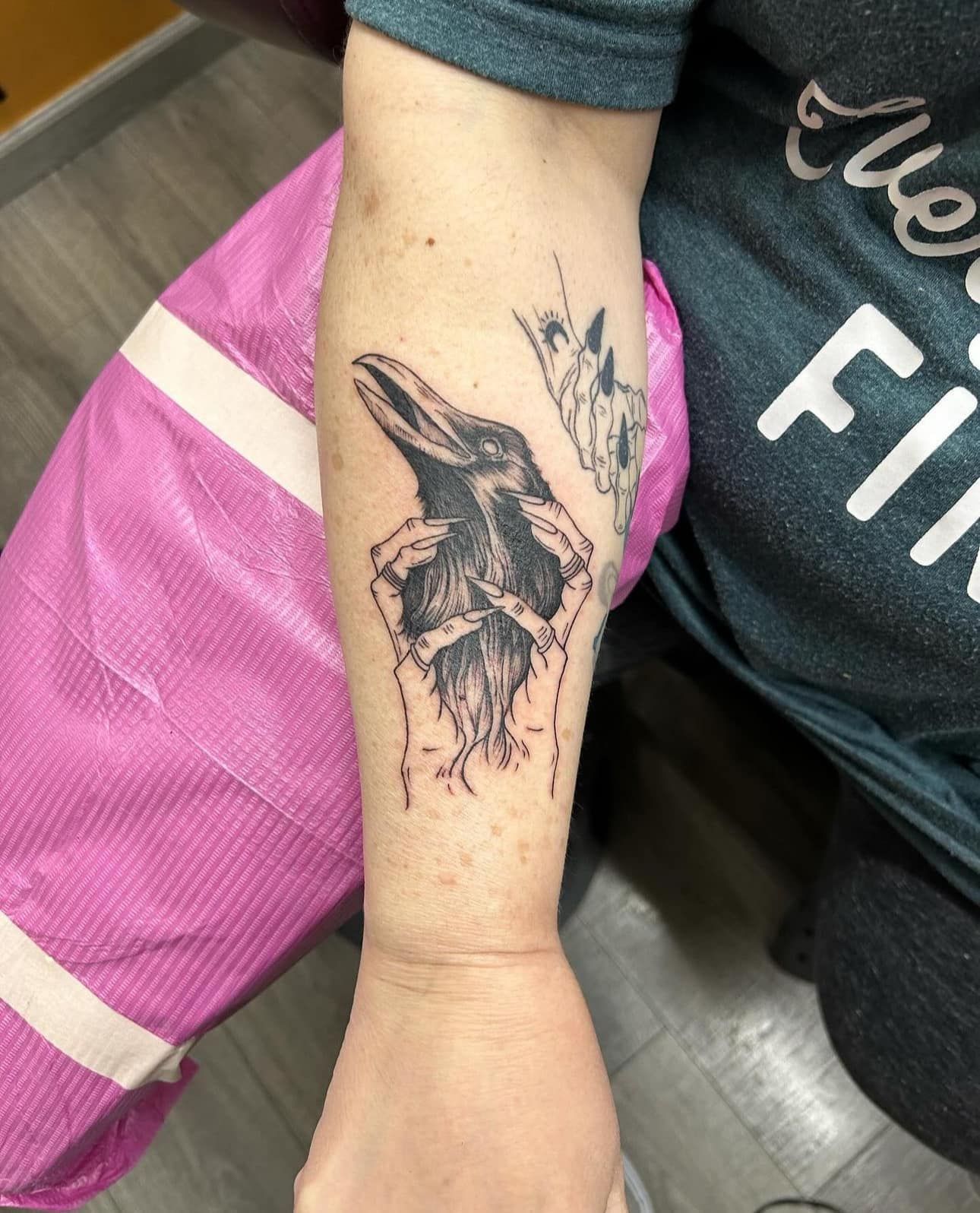 Forearm with a black bird tattoo on a pink chair, wearing a dark shirt and pink shorts