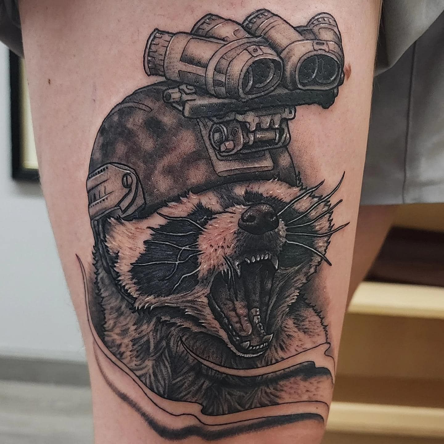 Black-and-gray tattoo of a roaring raccoon wearing a helmet with night-vision goggles on skin