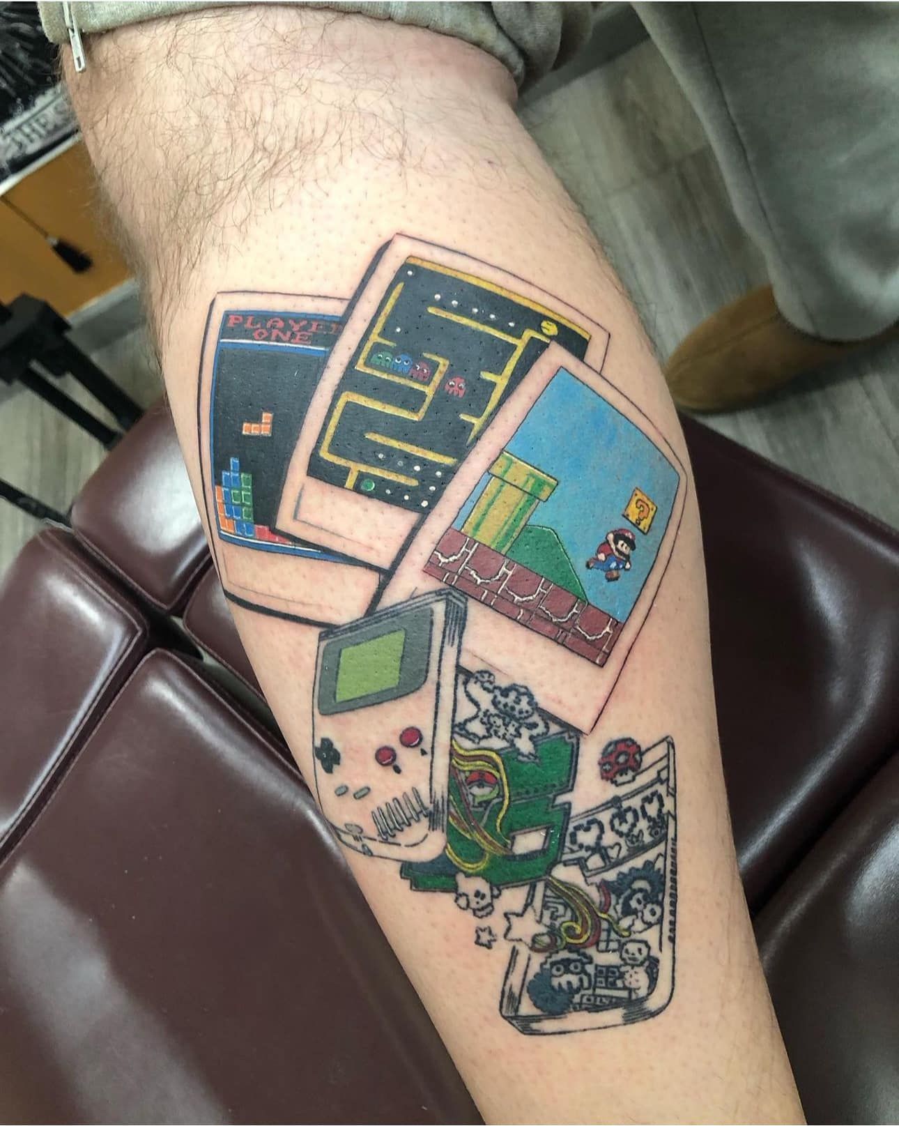 Colorful pixel-art tattoo on a person’s leg, showing three retro game screens and a green game controller.