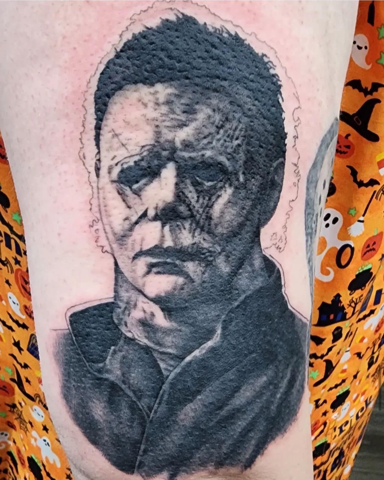 Black-and-gray tattoo portrait of a stern-faced man on skin, with a dark coat collar and orange patterned background