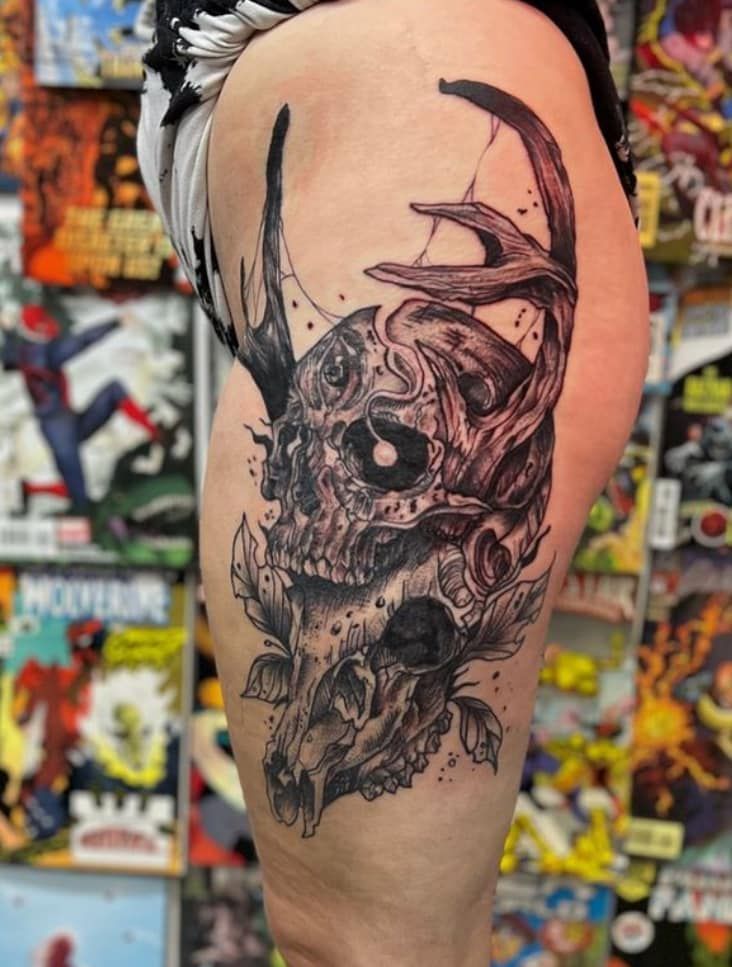Black-and-gray skull tattoo on a person’s upper thigh against a colorful comic-book background