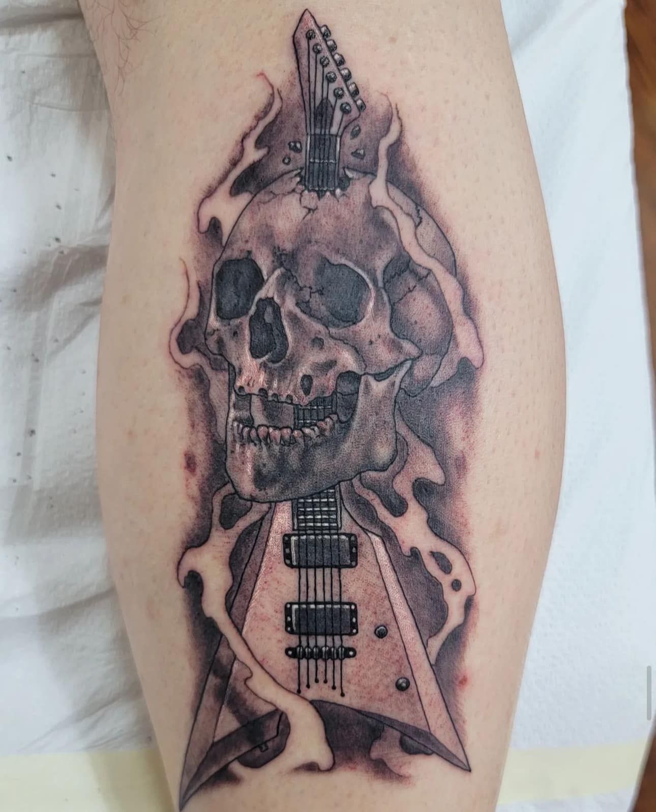 Skull-shaped black-and-gray guitar tattoo on a forearm with dripping details