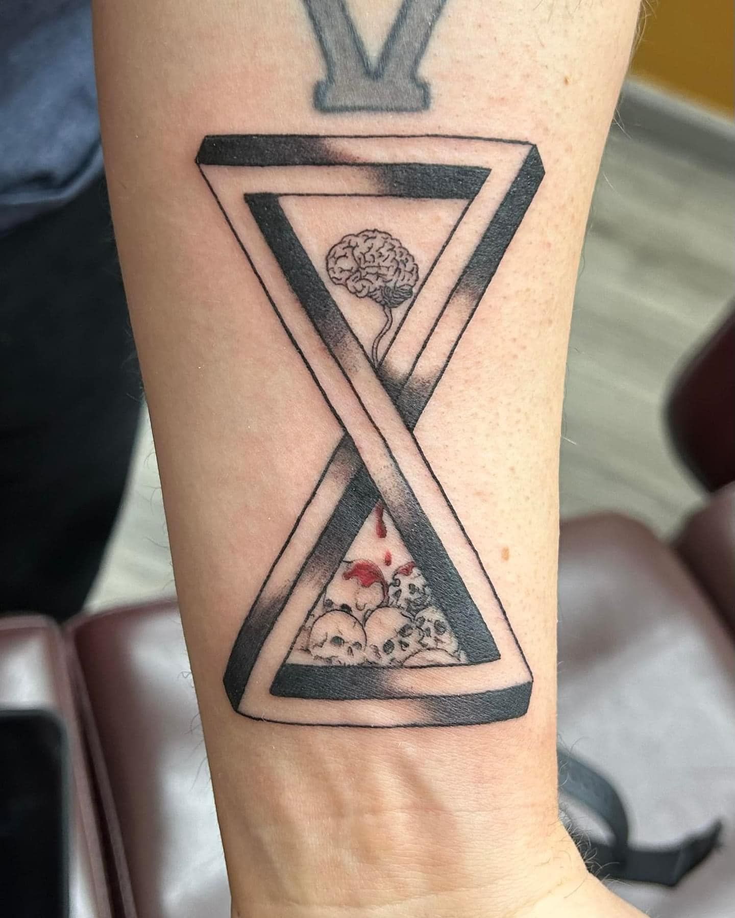Black hourglass tattoo with triangle frames and skull/floral details on a forearm.